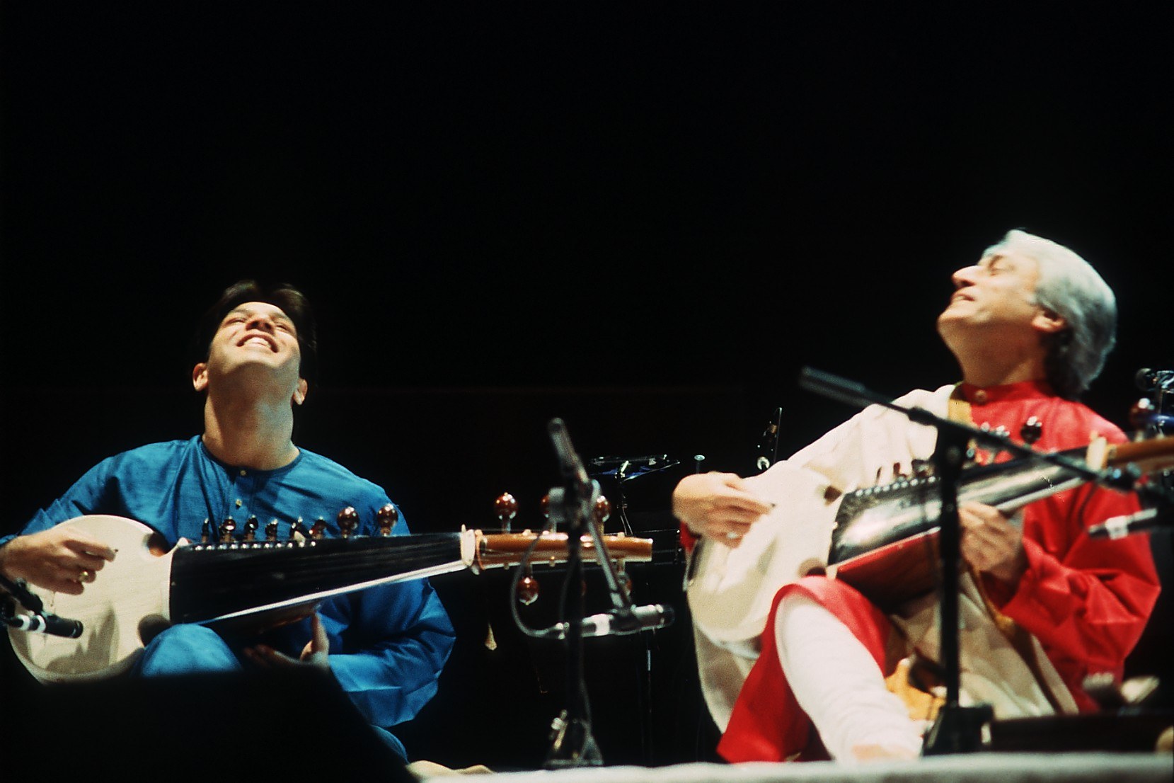 Two sarod players seated side by side on a concert stage.