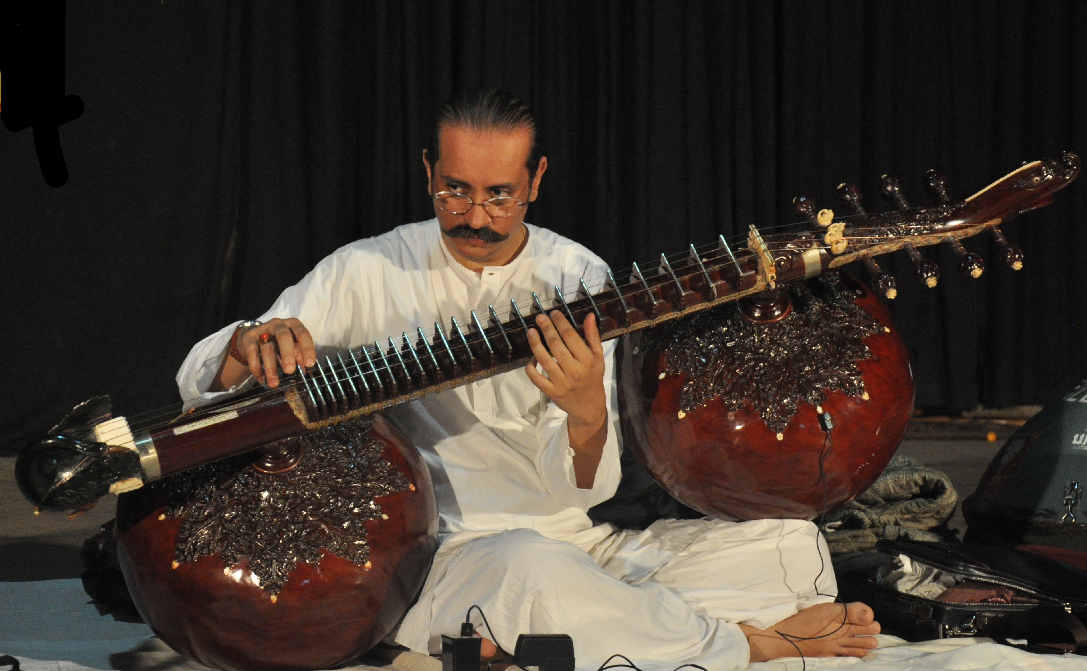 Bahauddin Dagar seated cross-legged playing a rudra veena, the instrument's two large gourds resting across his shoulder and knee.