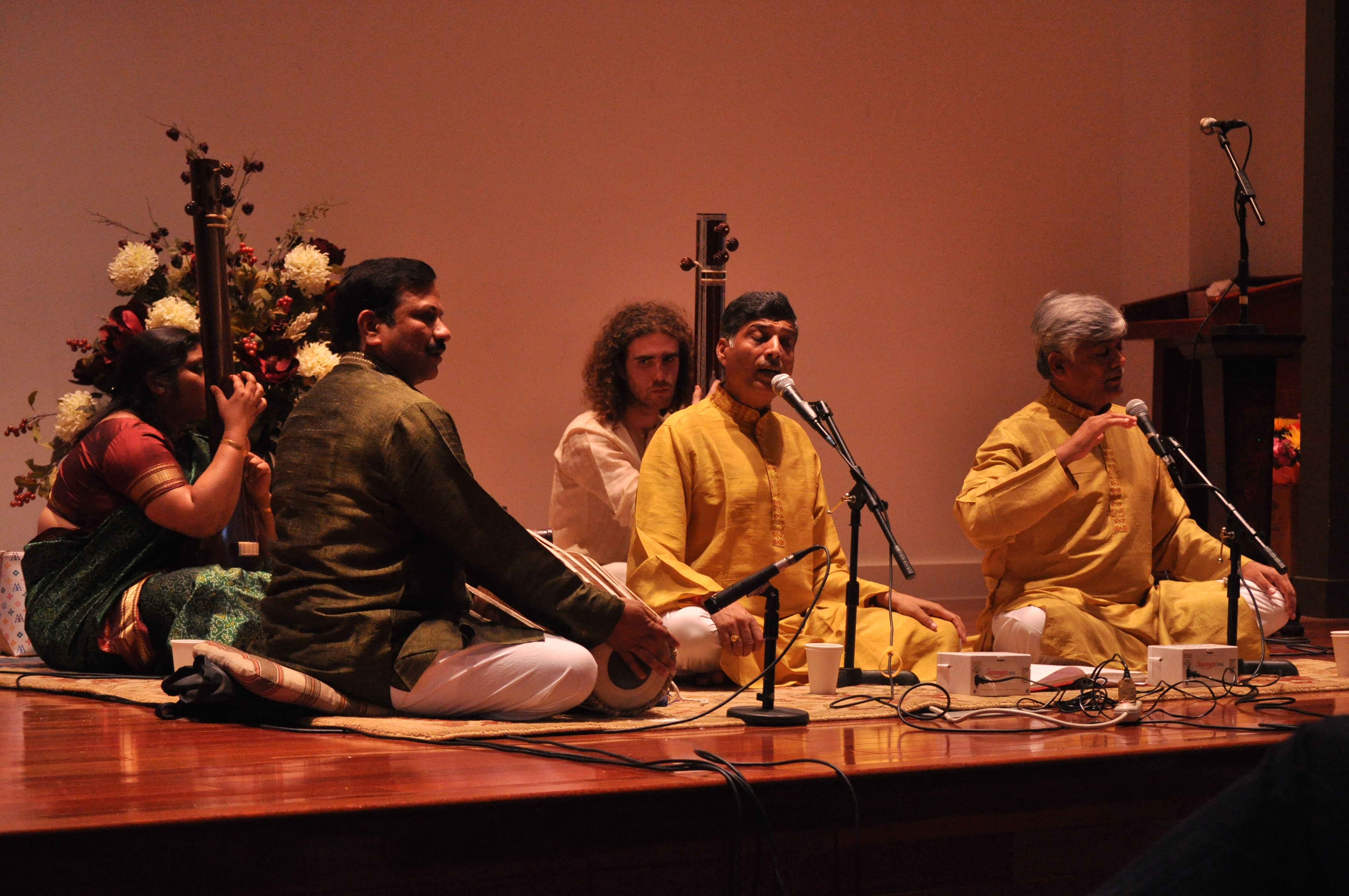 Two dhrupad singers seated on a low stage with tanpuras and a pakhawaj player beside them during a concert.