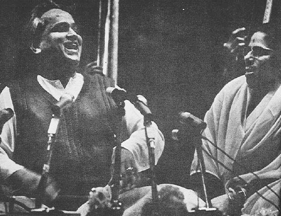 Kumar Gandharva laughing mid-performance beside Vasundhara Komkali, microphones between them.