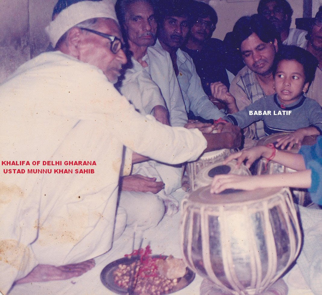 An elderly tabla master in a white cap and kurta leans forward to tie a red thread on the wrist of a small boy seated behind a pair of tabla, watched by a room of men.