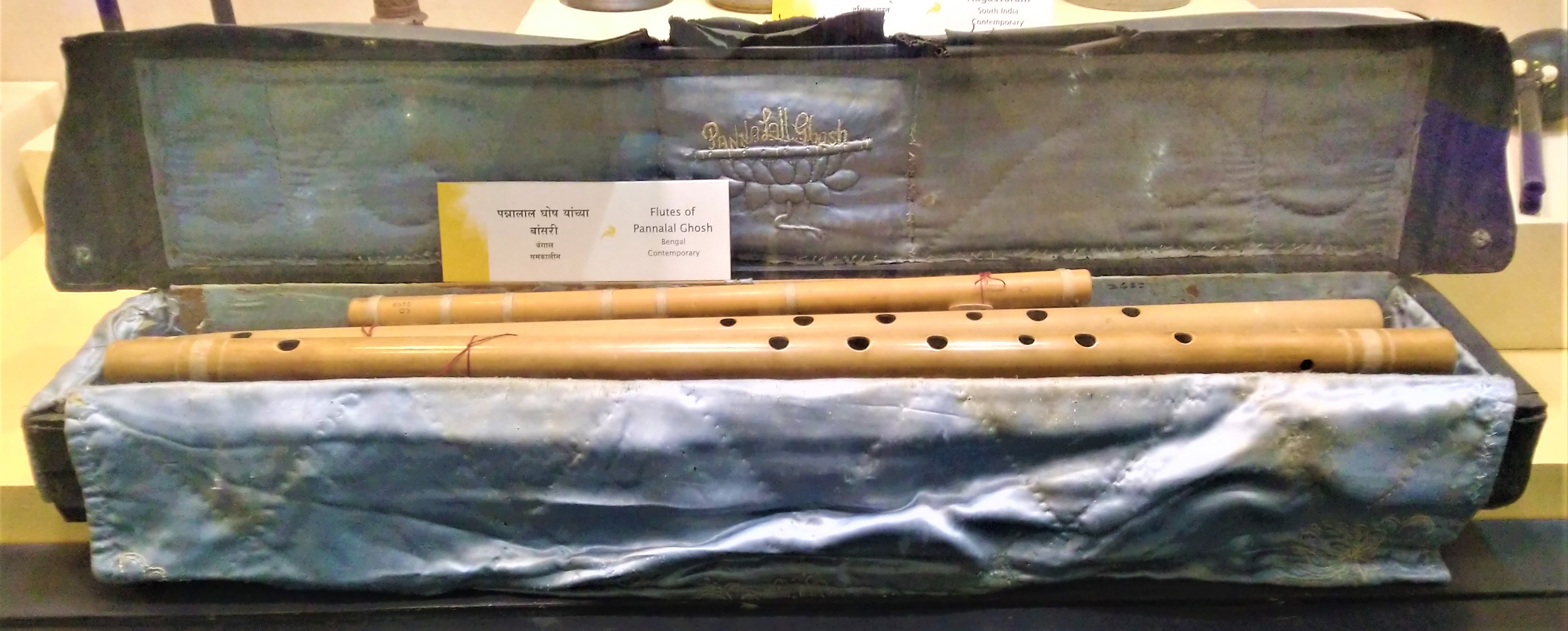 A set of bamboo flutes of varying lengths belonging to Pannalal Ghosh, displayed on his embroidered cloth flute bag in a museum case.