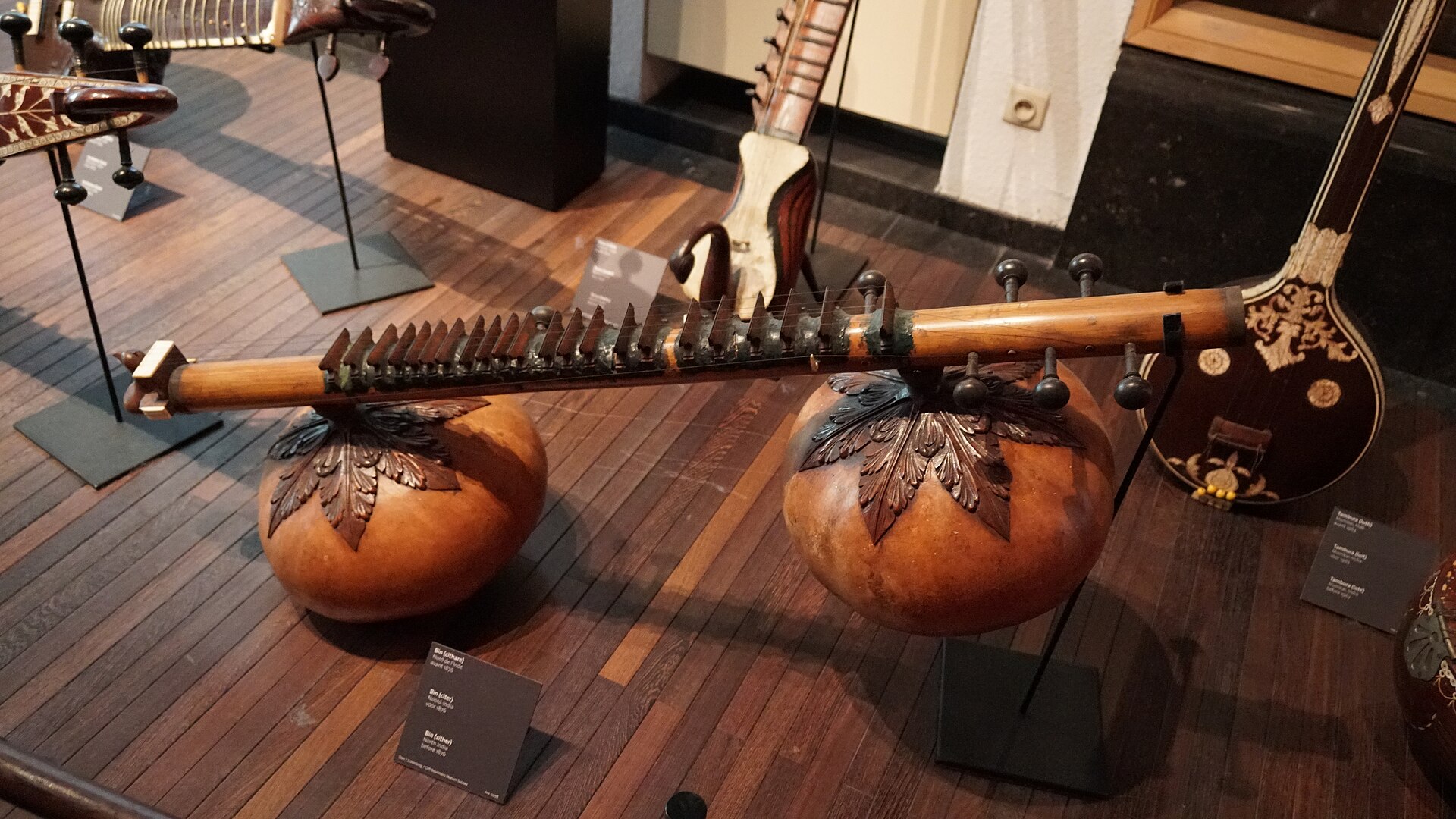 A rudra veena lying horizontally in a museum display, its two large gourds beneath a thick fretted tube.