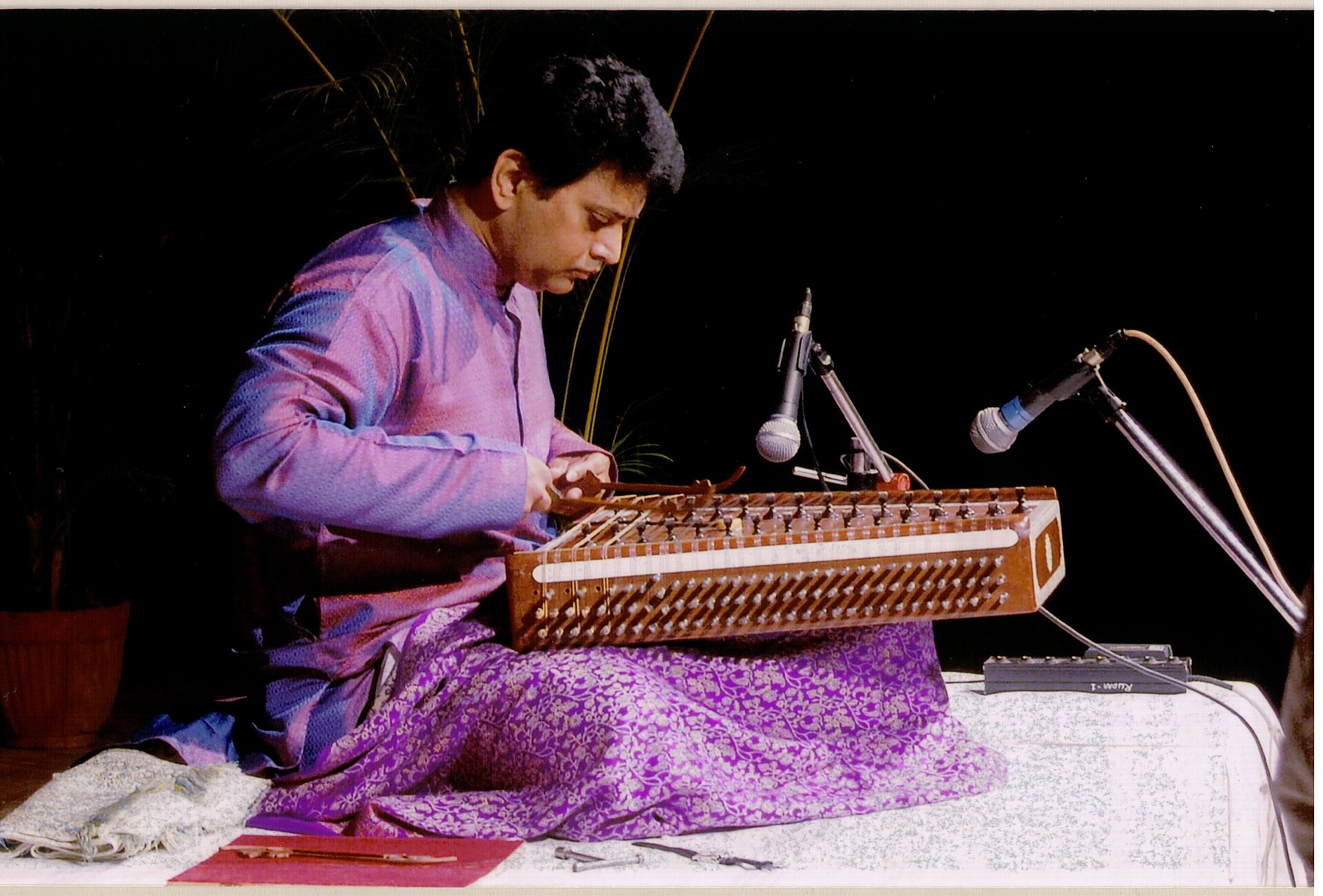A seated musician striking the strings of a trapezoidal santoor with two mallets.