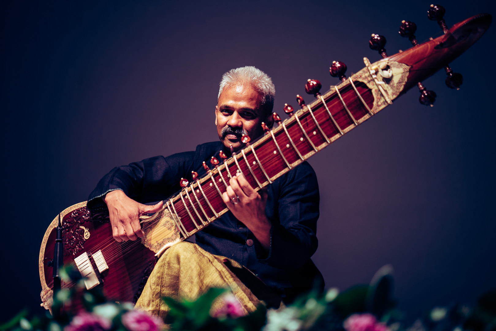 Seated musician playing a surbahar, a large long-necked plucked instrument.