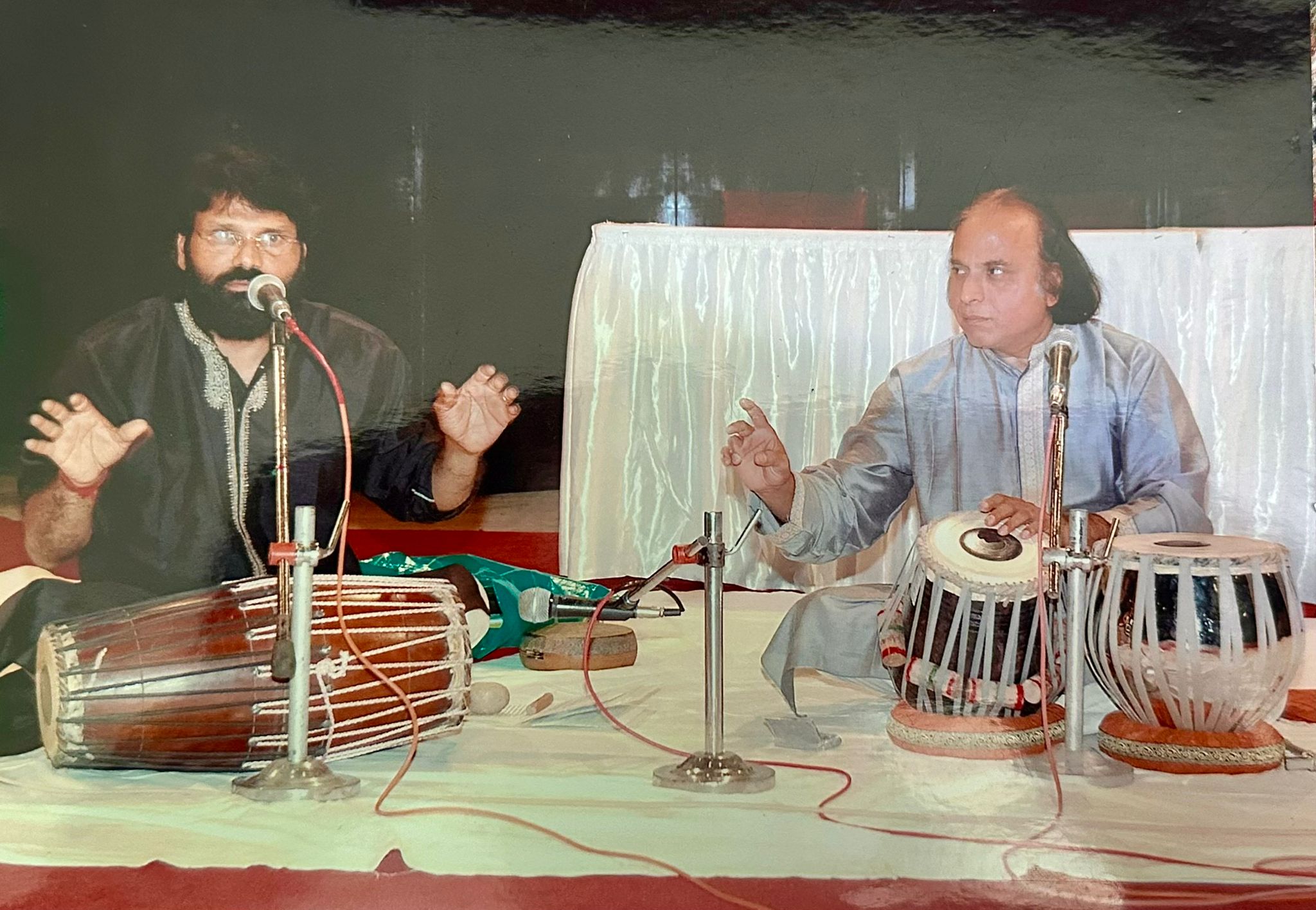 Suresh Talwalkar seated at the tabla on the right, speaking, with a mridangam player on the left.